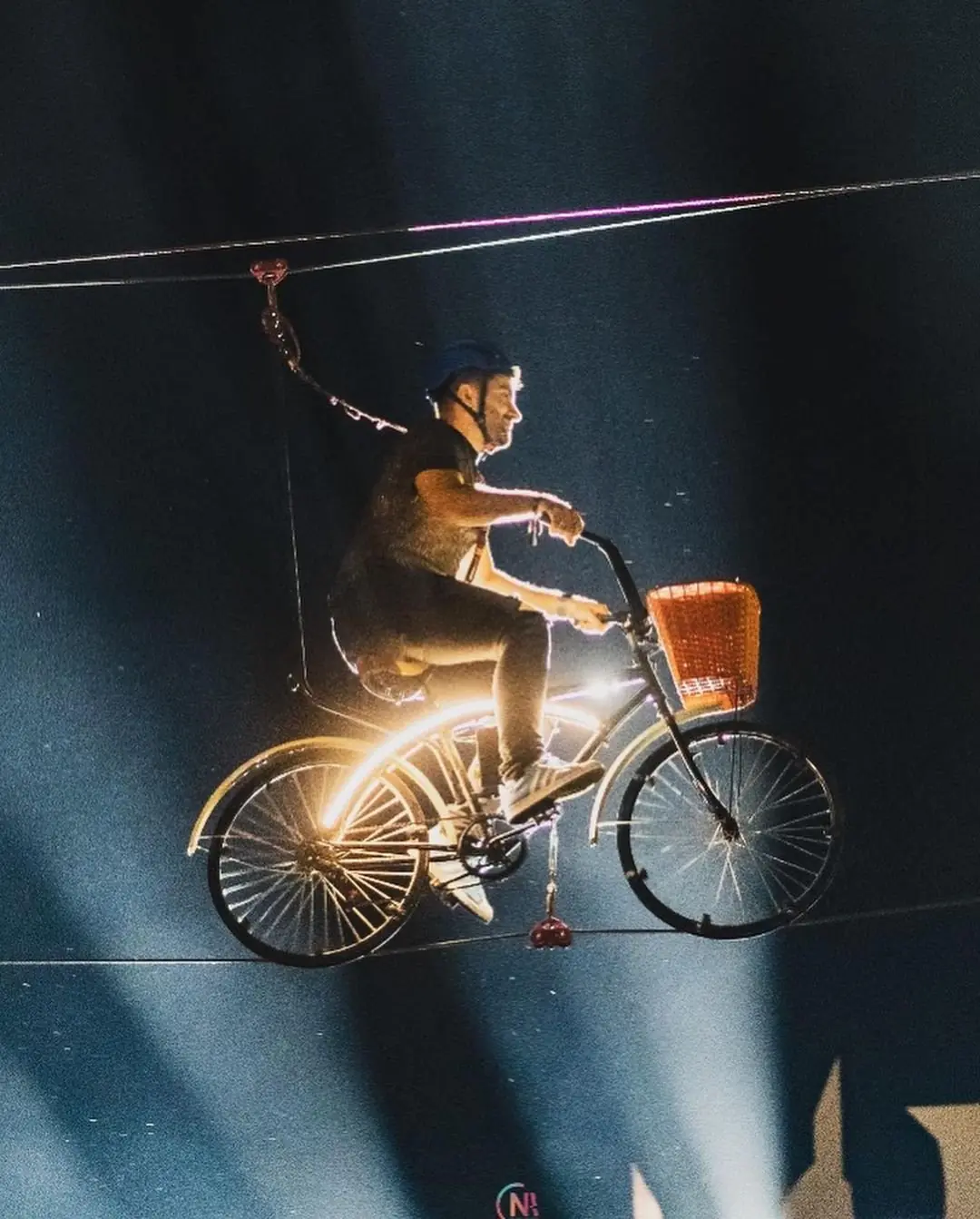 Pessoa pedalando na Sky Bike sobre um cabo suspenso, iluminada durante apresentação